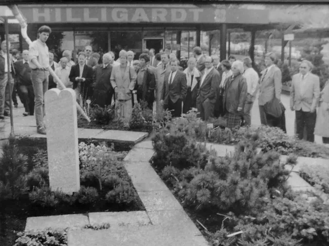 Schwarzweißfoto 1989: Gruppe von Menschen steht andächtig vor einem frisch bepflanzten Grab, im Hintergrund der Schriftzug „HILLIGARDT“ über dem Geschäft
