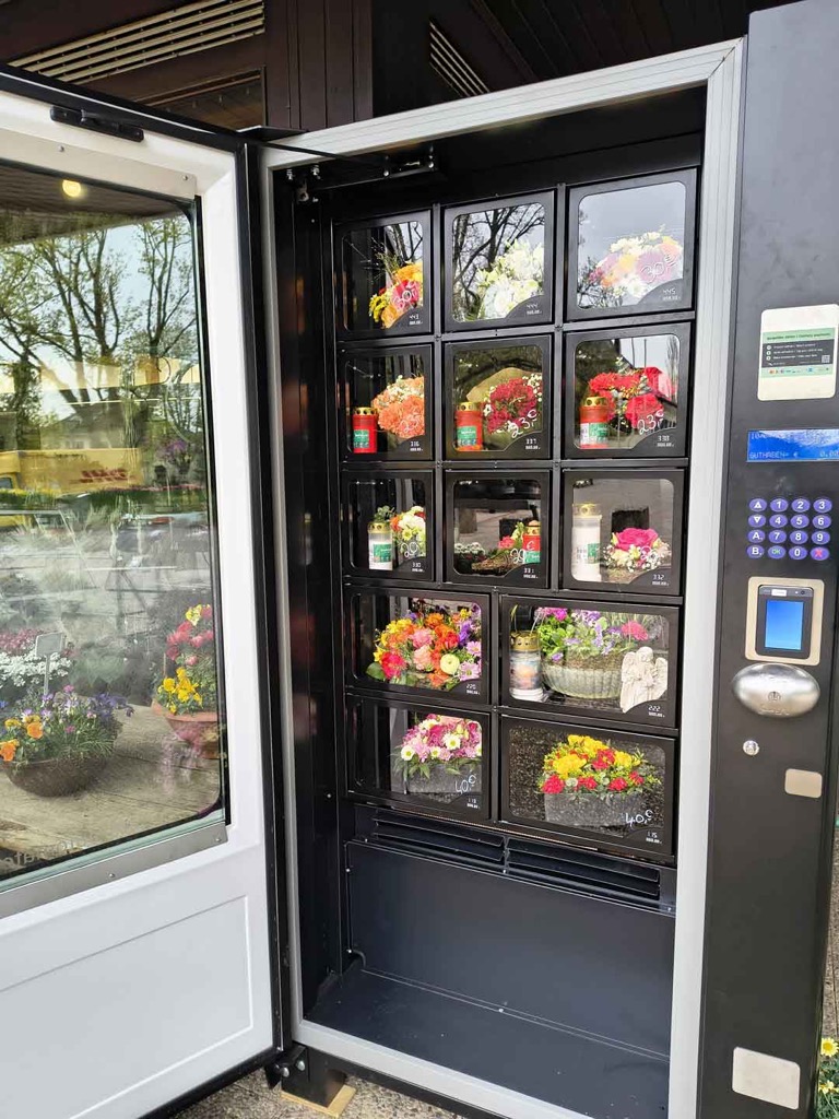 Geöffneter Blumenautomat mit beleuchteten Fächern voller Sträuße und Grableuchten