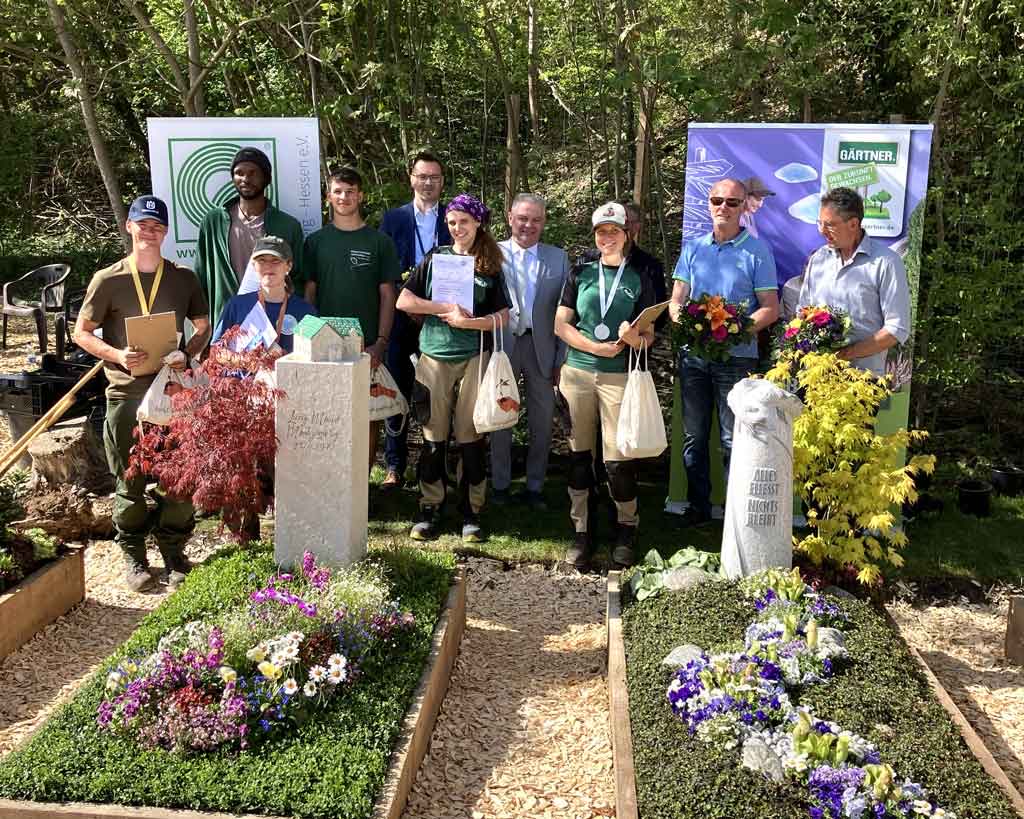 Preisverleihung im Freien: Gruppe von etwa zehn Personen hinter zwei frisch gestalteten Gräbern mit Blumenschmuck und Urkunden in der Hand
