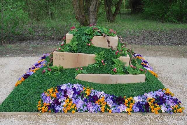 Terrassierte Grabbepflanzung aus Rasen und Moos in Pyramidenform mit Natursteinstufen, Inschrift „FAMILIE RHEIN“, Farnbeete, umrandet von violetten und orangen Stiefmütterchen