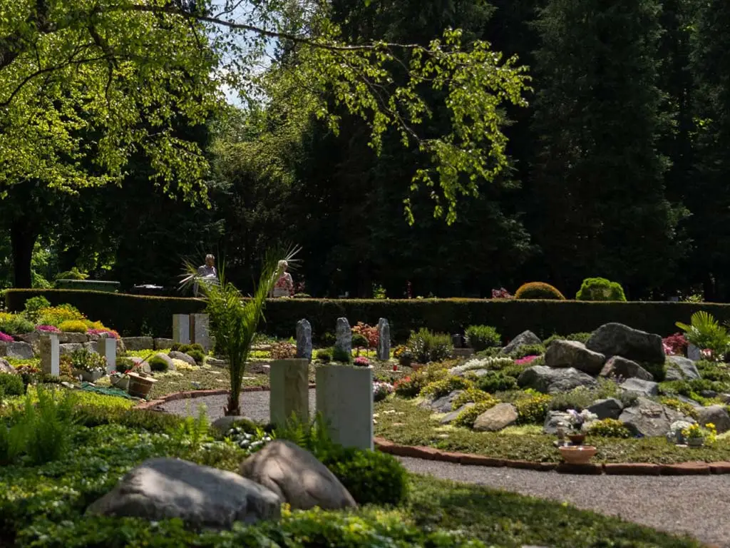 Weitläufige Friedhofsanlage mit Kiesweg, Steingarten-Gräbern mit niedrigen Grabsteinen und altem Baumbestand