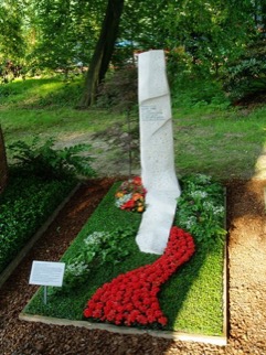 Schlanke, schräg aufragende weiße Steinstele auf Rasenfläche, rote Blumenkaskade an der Basis, kleiner Kranz