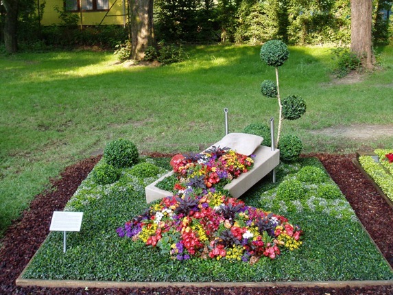 Grabgestaltung als Bett-Skulptur aus hellem Stein mit Kissen, bunte Blumen als „Decke“ drapiert, Buchsbaumkugeln und Formschnitt-Bäumchen