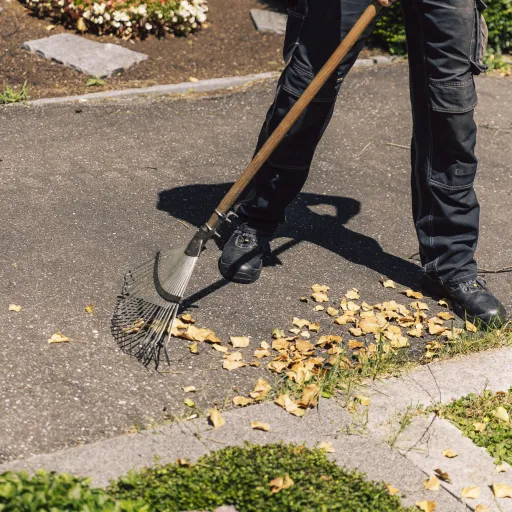 Person recht Laub von einem Weg neben einem Blumenbeet, nur Beine und Schuhe im Bild