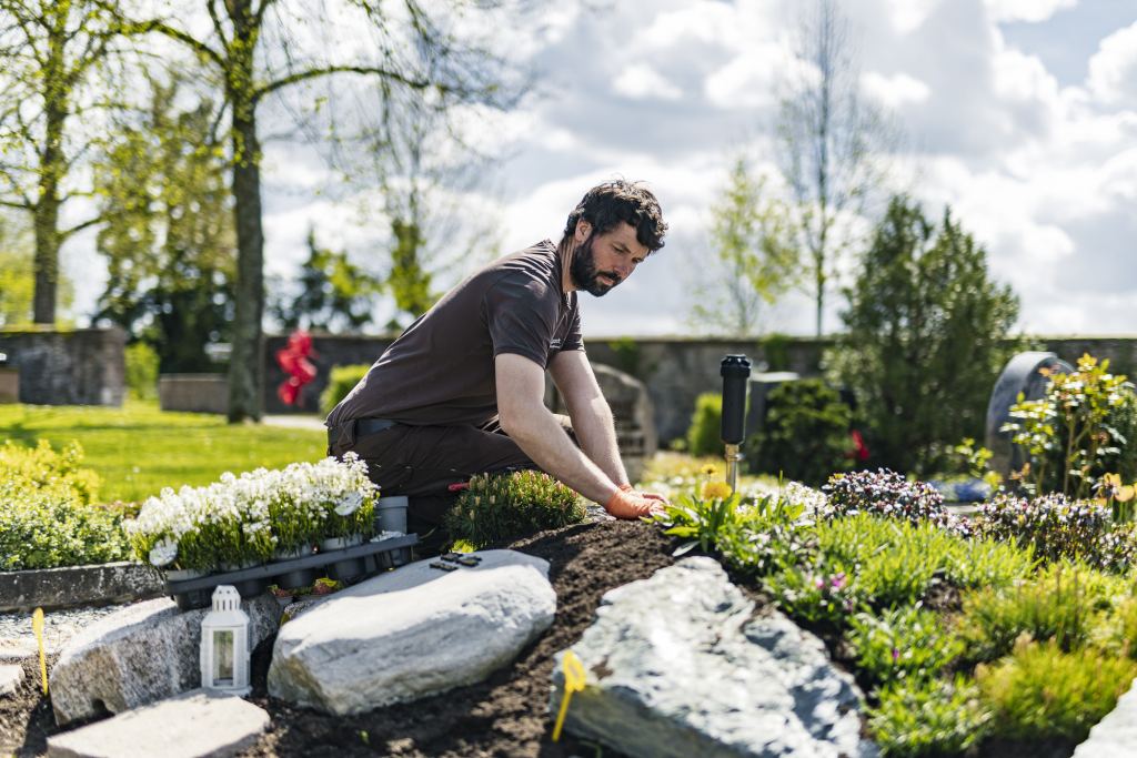 Gärtner mit dunklem Bart in brauner Arbeitskleidung kniet an einem frisch bepflanzten Grab und drückt Pflanzen in die Erde, vorne große helle Natursteine, links Pflanzkisten mit weißen Blumen