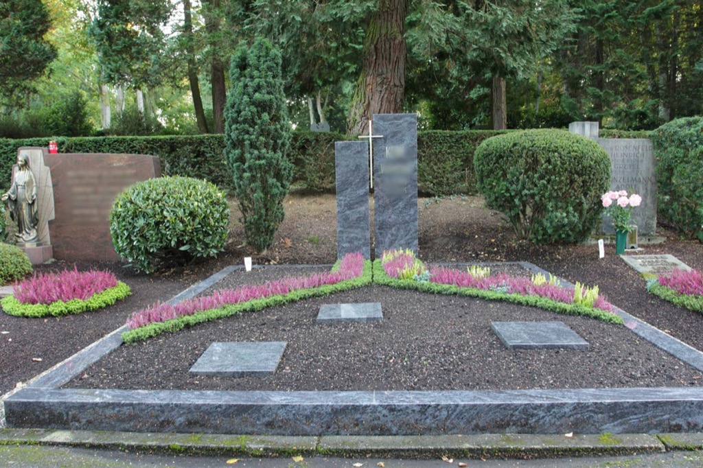 Breites Familiengrab mit dunklem Kreuz-Grabstein, Buchsbaumkugeln, dreieckige Beete mit pinker Erika, Engelsstatue links