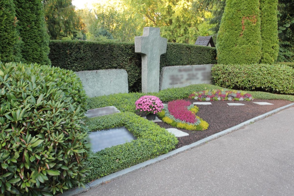 Grab mit steinernem Kreuz, dichte Buchseinfassung, pinke Chrysanthemen und rote Sedum-Blüten im Beet