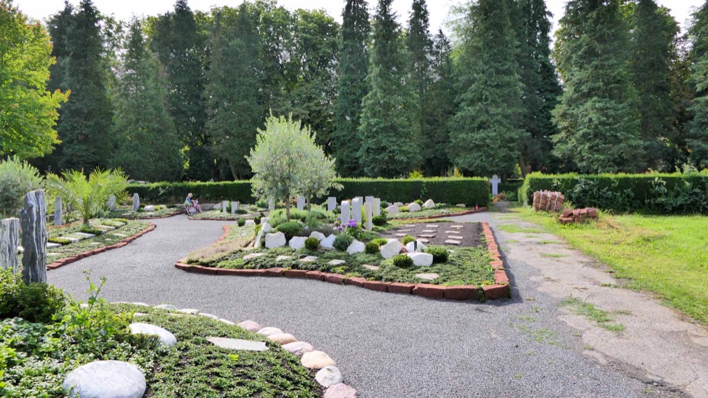 Geschwungener Kiesweg durch ein Gräberfeld mit schlanken weißen Grabstelen, jungem Ölbaum, Findlingen und niedrigen Hecken