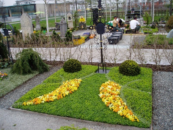 Rechteckiges Rasengrab mit zwei geschwungenen Bändern aus gelben und orangen Blüten, schmiedeeisernes Ornament-Element mittig, zwei Buchsbaumkugeln