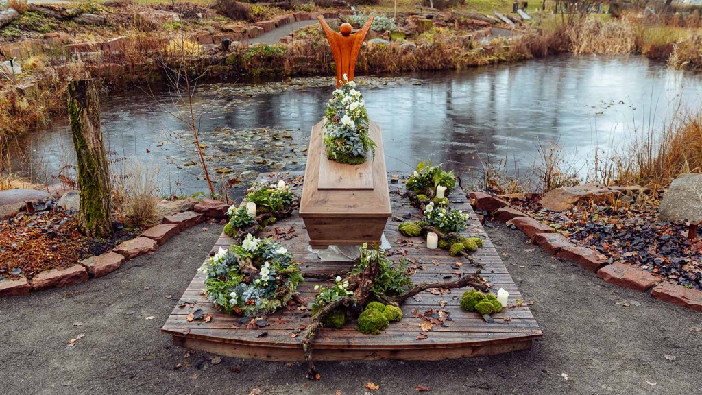 Sargschmuck aus Christrosen, Farnen, Moos und Zapfen auf einem hellen Holzsarg, aufgebahrt auf einem Holzsteg an einem winterlichen Teich, daneben Kränze, Wurzelholz und Grablichter