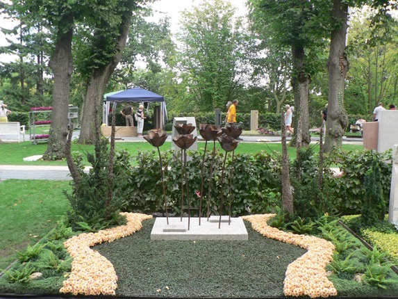 Reihe stilisierter Mohnblüten-Skulpturen aus Metall auf weißer Steinplatte, umrandet von hellen Kieselsteinen auf dunklem Rasen