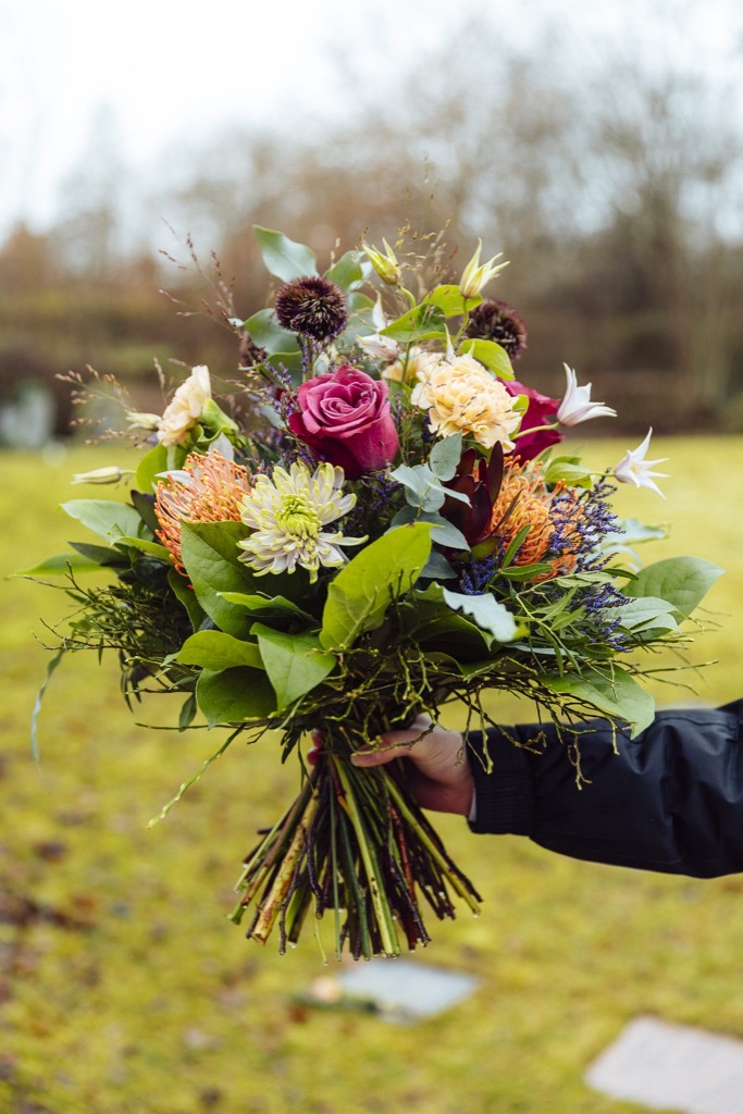 Eine Hand hält einen gebundenen Strauß mit pinken Rosen, hellen Chrysanthemen, orangen Blüten und Eukalyptus, im Hintergrund eine Wiese