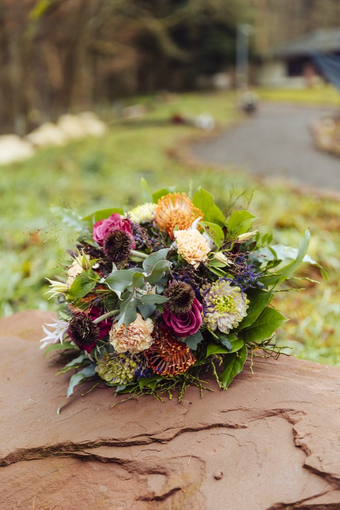 Runder Strauß mit pinken Rosen, apricotfarbenen Nelken, orangen Blüten und dunklen Skabiosen auf einer Sandsteinplatte im Freien