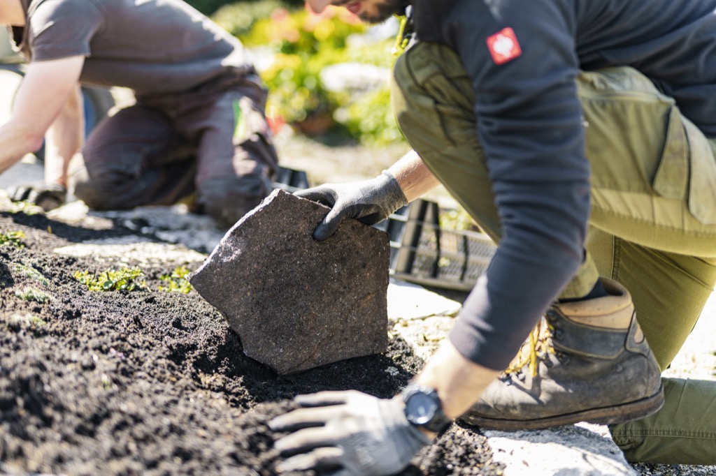 Zwei Mitarbeitende in Arbeitskleidung knien auf frisch aufgeschütteter Erde und setzen eine Natursteinplatte, die Gesichter sind nicht im Bild