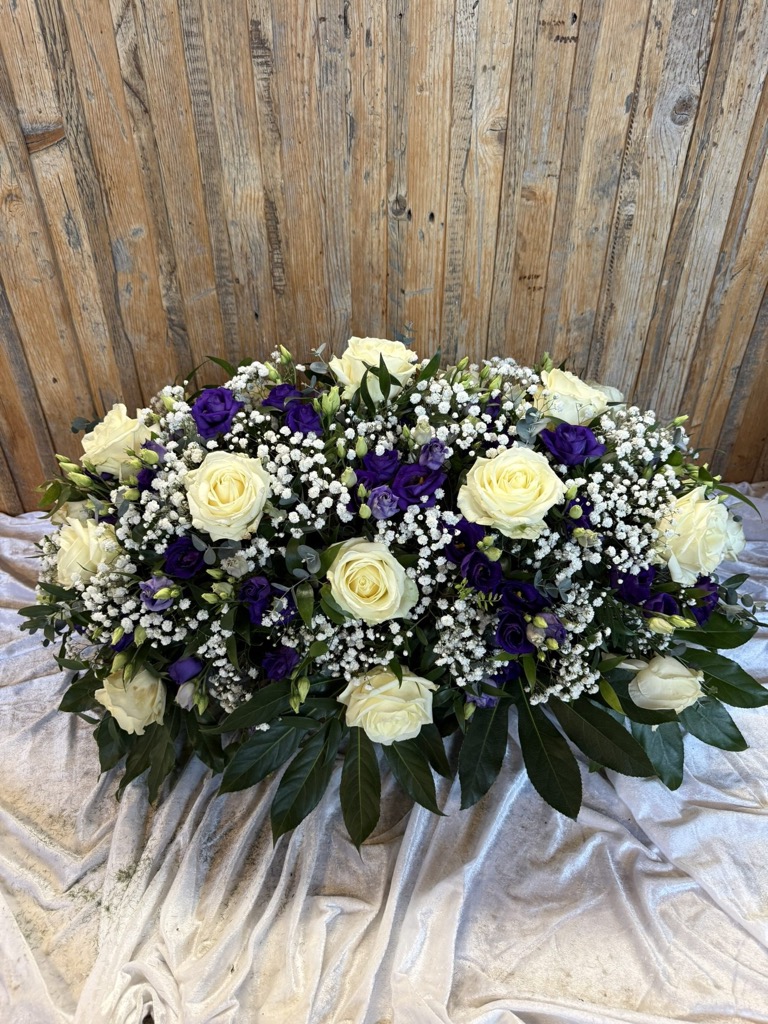 Längliches Trauergesteck aus weißen Rosen, Schleierkraut und violetten Blüten auf weißem Tuch