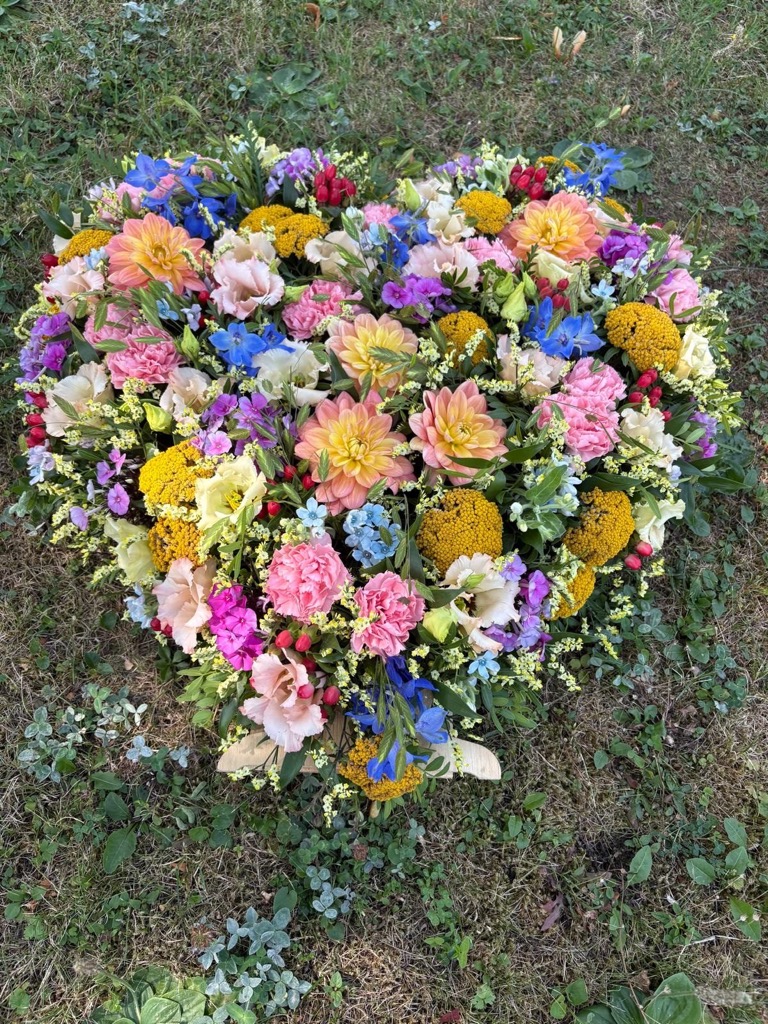 Großes Herz aus bunten Blüten in Rosa, Gelb, Blau und Weiß, auf einer Wiese liegend