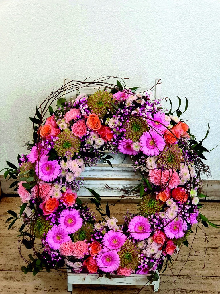 Trauerkranz mit pinken Gerbera, orangen Rosen, Nelken und Zweigen auf einem weißen Holzständer