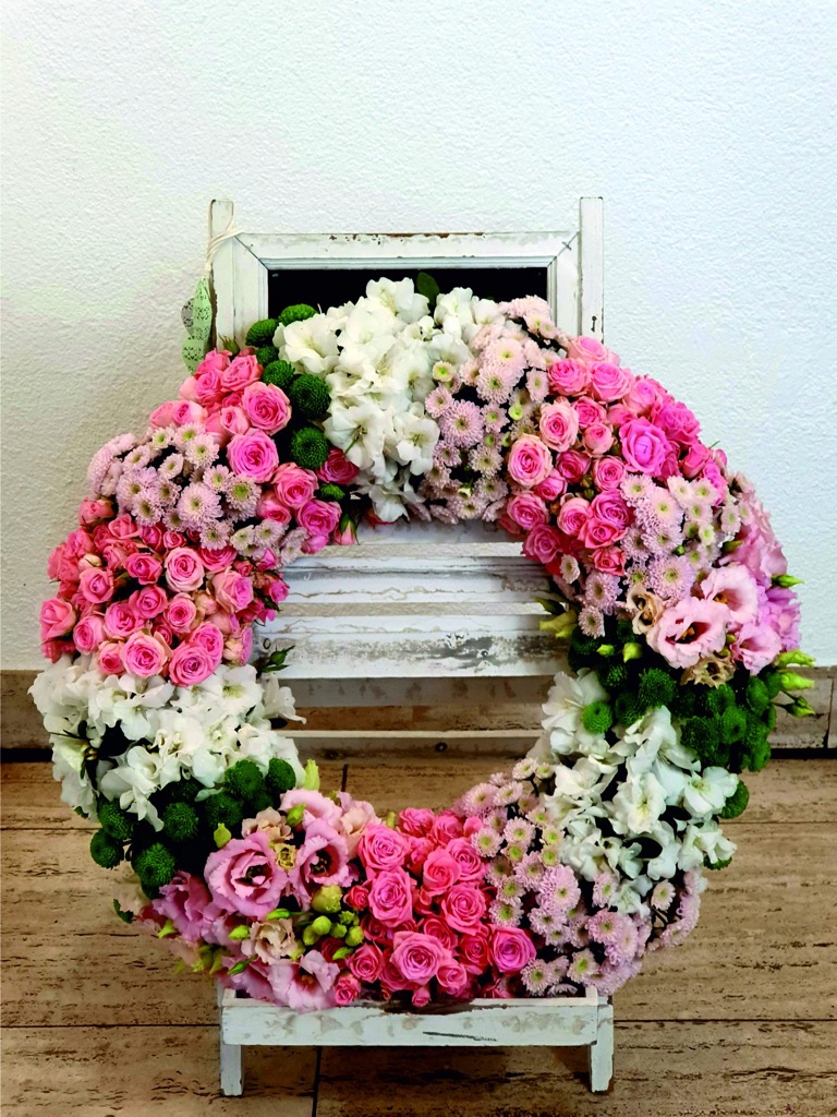 Trauerkranz in Abschnitten aus rosa Rosen, weißen Blüten und grünen Chrysanthemen auf einem weißen Holzständer