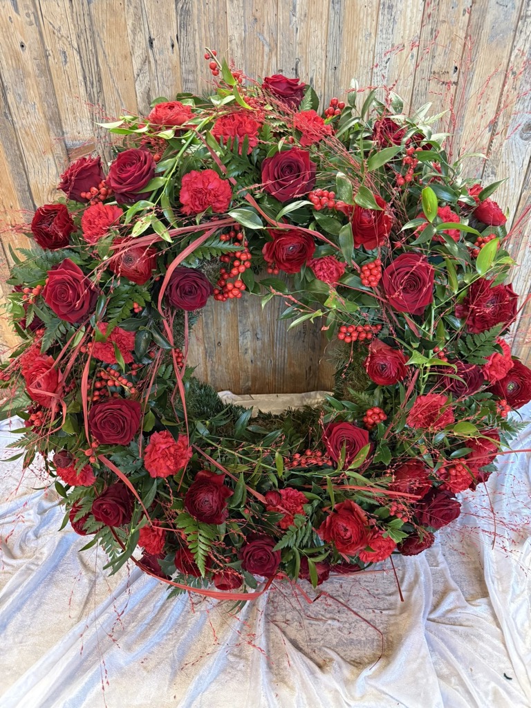 Runder Trauerkranz aus roten Rosen, Nelken, roten Beeren und Zweigen auf weißem Tuch vor einer Holzwand