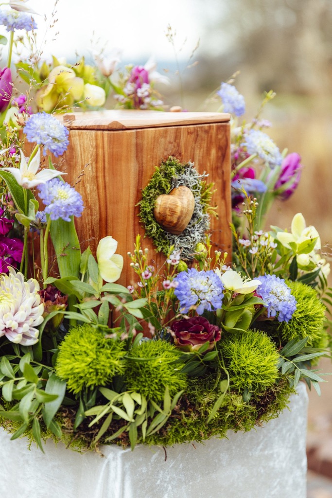 Holzurne mit einem Herz aus Moos, umgeben von einem Kranz aus Wiesenblumen, Skabiosen und grünen Nelken
